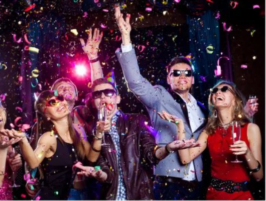 A group of friends celebrating a birthday with colorful confetti, wearing party hats and sunglasses while holding glasses of champagne.