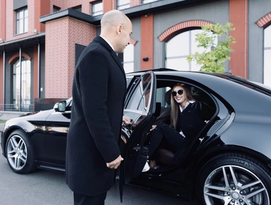 A professional chauffeur holding the door for a businesswoman as she exits a black luxury sedan.