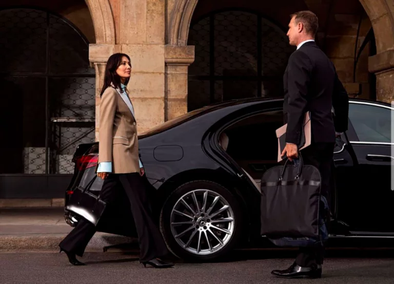 A professional chauffeur holding luggage for a businesswoman as she walks toward a luxury sedan
