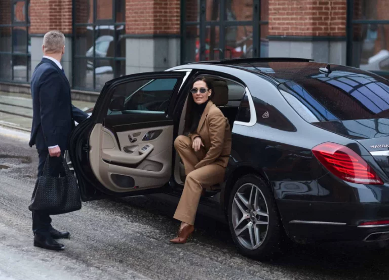 A professional chauffeur holding the door for a businesswoman as she exits a black luxury sedan.