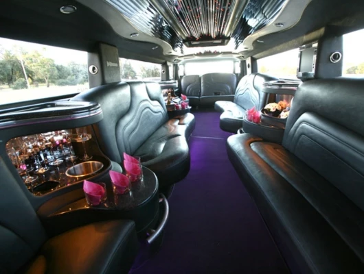 A front three-quarter view of a black Hummer stretch limousine featuring chrome grille accents, chrome side mirrors, and custom multi-spoke wheels. The vehicle is parked on a dirt lot with other cars and green trees visible in the background under a clear blue sky.