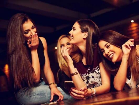 A group of four young women laughing and smiling together during a night out at a dimly lit venue, with one holding a smartphone.