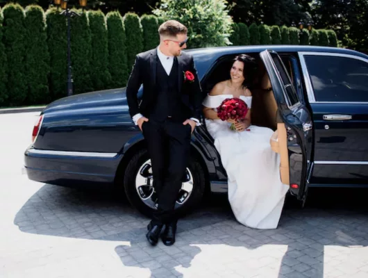 A bride holding a red bouquet sitting in the back of a luxury car while the groom in a black suit leans against the exterior.